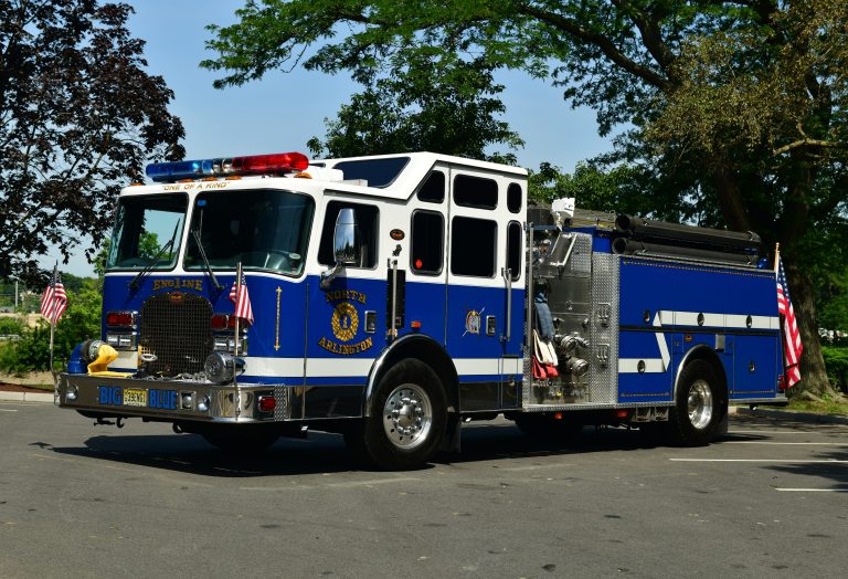 A blue and white fire truck with American flags on the front and sides is parked on a street, surrounded by green trees. The truck is labeled "North Branford" and has "BIG BLUE" on its front license plate.