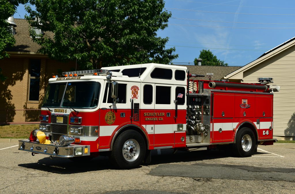 A red and white fire truck labeled "SCHUYLER ENGINE 6" is parked on a paved lot near buildings and trees on a sunny day.