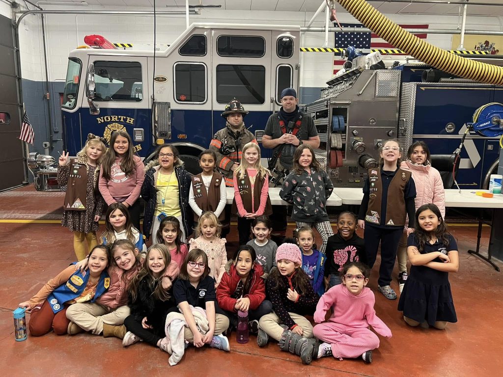 A group of Girl Scouts posing with two firefighters in uniform in front of a fire truck inside a fire station. The girls are smiling and wearing their vests or sashes, and some are sitting while others are standing.