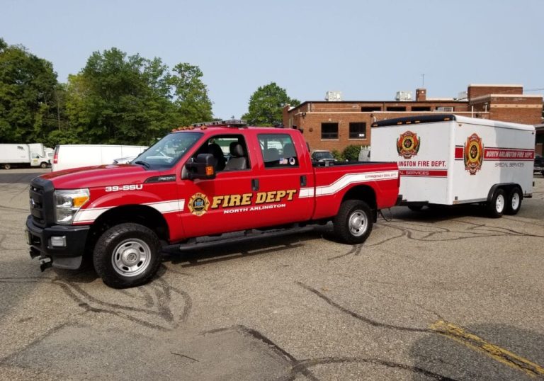 A red North Arlington Fire Department pickup truck with white stripes is towing a matching fire department trailer in a parking lot, with trees and brick buildings in the background.
