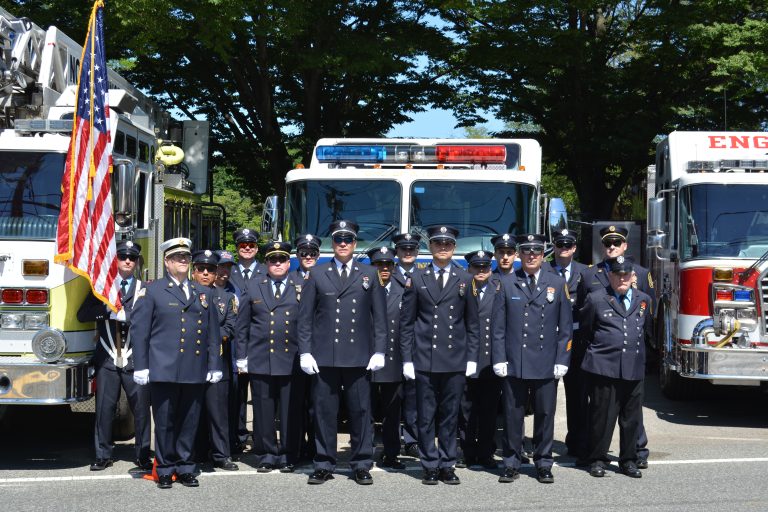 A group of uniformed firefighters stands in front of two fire trucks, one of them holding an American flag. Trees provide shade in the background on a sunny day.
