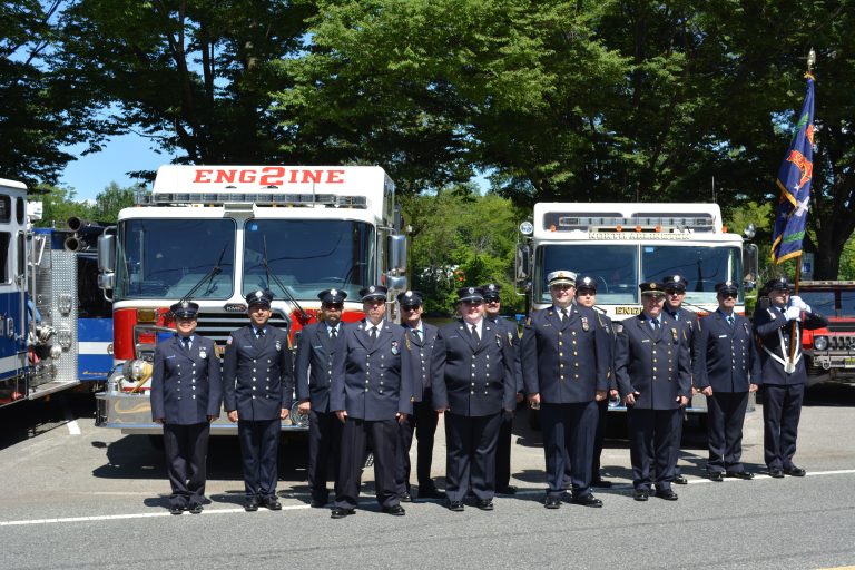 A group of firefighters in uniform stand in front of two fire trucks labeled "Engine 2" during the daytime, with trees and parked vehicles in the background.