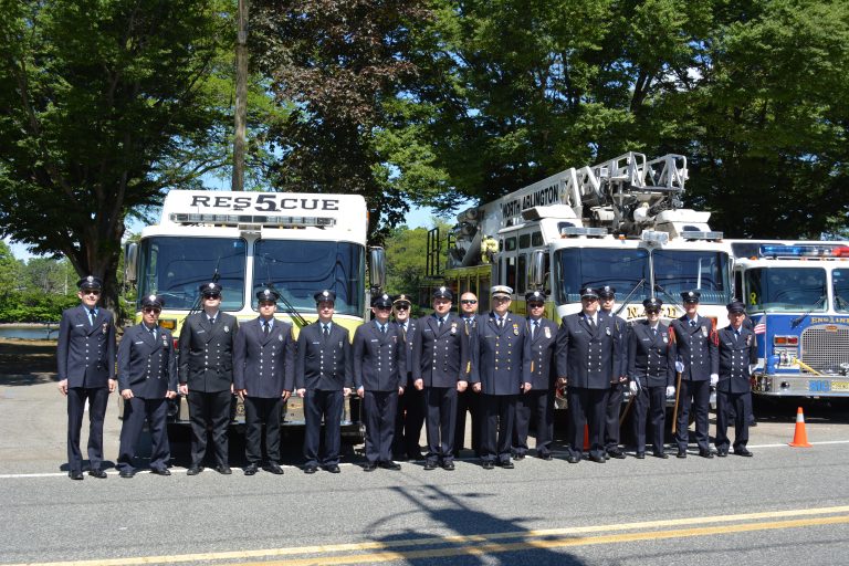 A group of firefighters in dress uniforms stand in a row in front of rescue and fire trucks parked on a sunny street, with trees in the background.