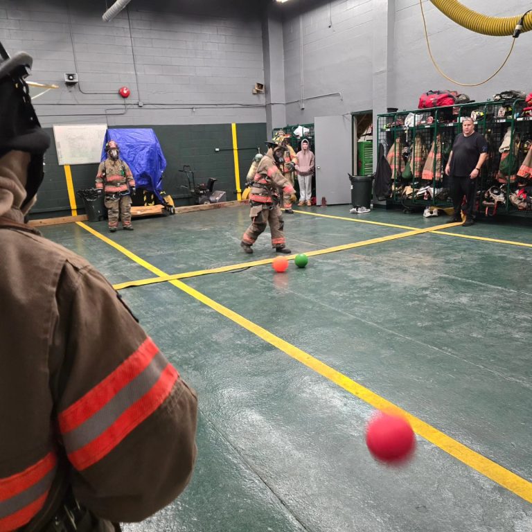 Firefighters in full gear play dodgeball with red and green balls inside a fire station. Several people watch near lockers with equipment, and the floor has yellow boundary lines.