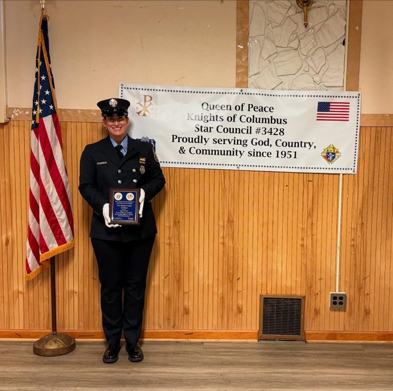 A smiling uniformed firefighter holds a plaque while standing next to an American flag and a banner that reads, "Queen of Peace Knights of Columbus Star Council #3428 Proudly serving God, Country, & Community since 1951.