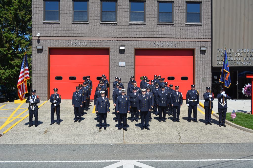 A group of uniformed firefighters stand in formation in front of two bright red garage doors at a fire station, with American flags and a memorial sign visible on the building.
