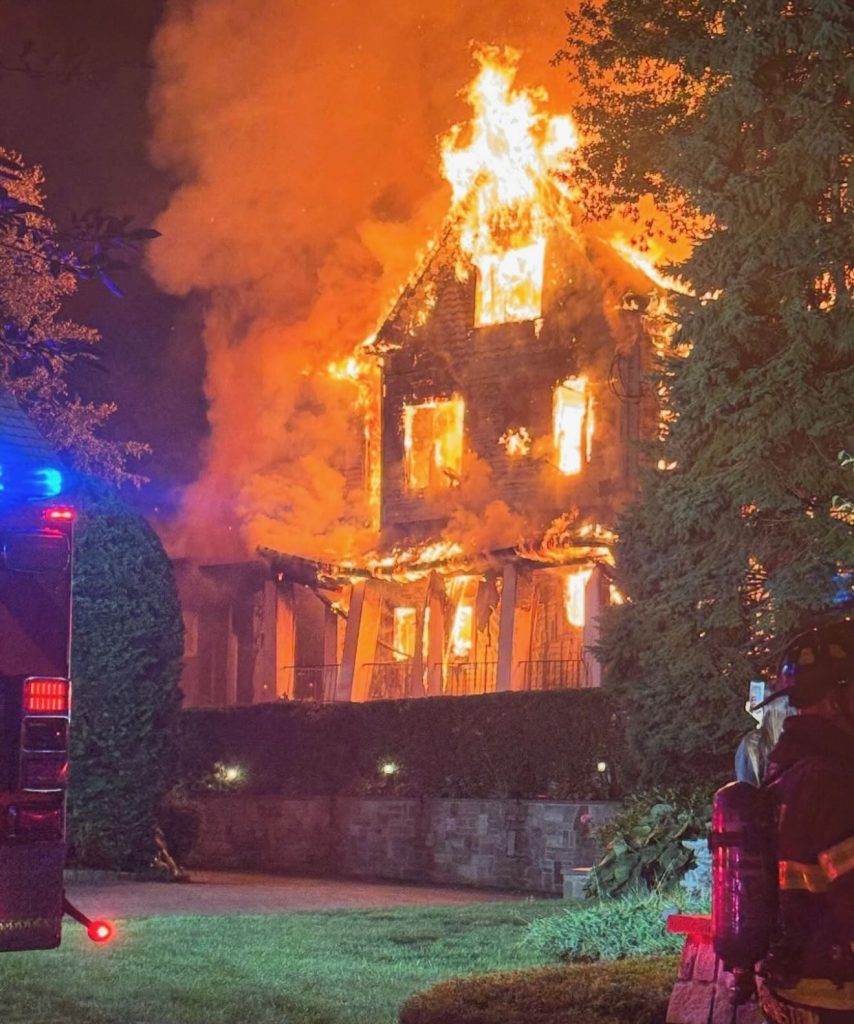 A large house is engulfed in bright orange flames at night. Smoke billows from the roof, and firefighters with helmets stand nearby as emergency vehicle lights flash on the scene.