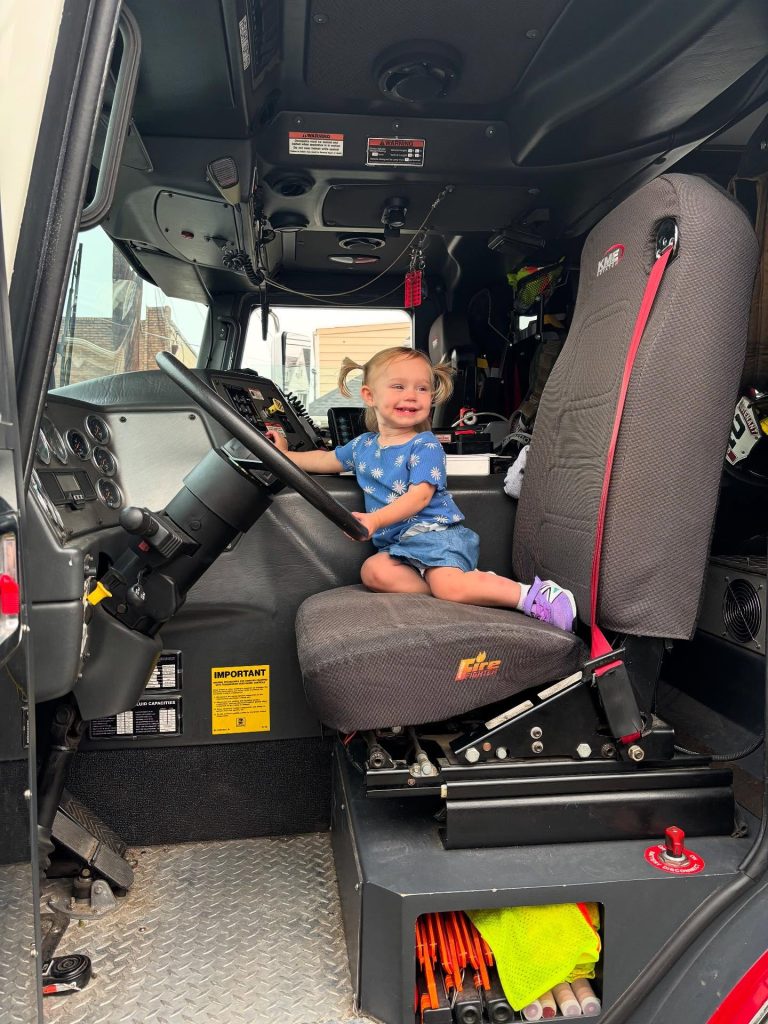 A smiling toddler in a blue dress and purple shoes sits in the driver’s seat of a large truck, holding the steering wheel and looking at the camera. The truck’s interior and dashboard are visible.