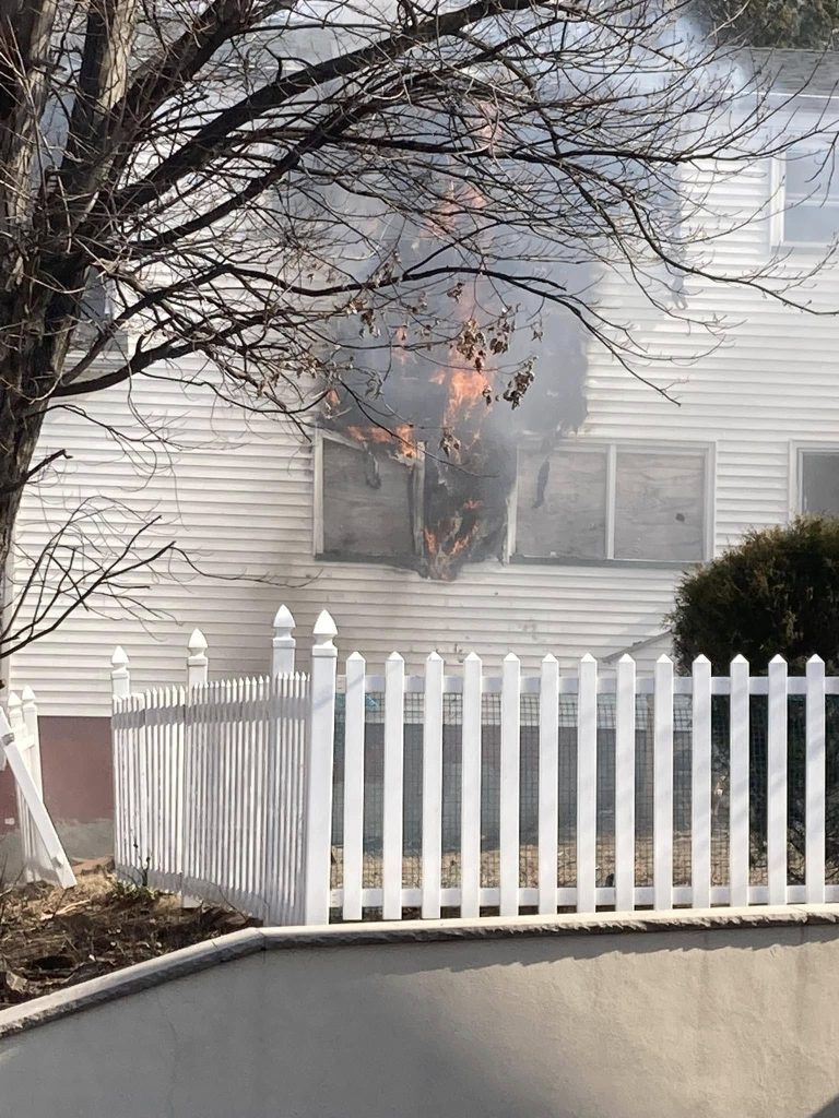 A fire blazes through a boarded-up ground floor window of a white house behind a white picket fence, with smoke rising and tree branches visible in the foreground.