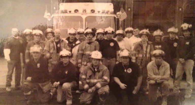 A sepia-toned photo of a group of firefighters in uniform and helmets posing in front of a fire truck with two American flags attached to it. Some are standing while others are kneeling in front.