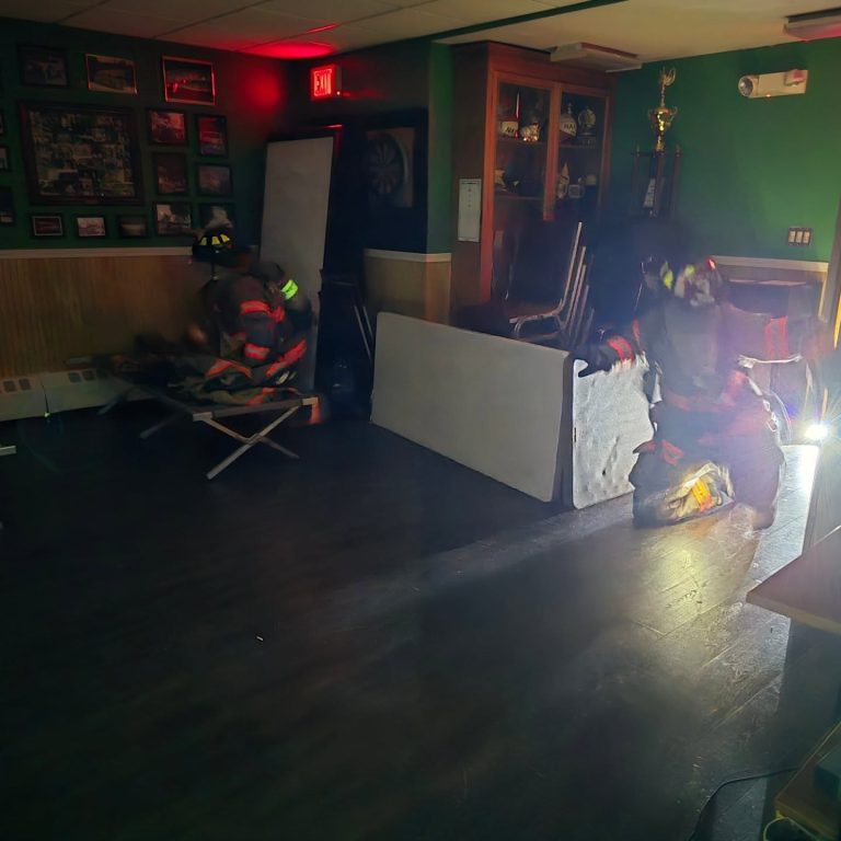 Two firefighters in full gear move through a dimly lit room with green walls, wooden furniture, and trophies. One kneels, shining a flashlight, while the other searches near a wall covered in photos.