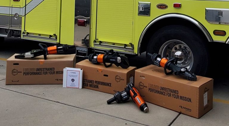 Three hydraulic rescue tools, also known as “Jaws of Life,” are displayed on cardboard boxes next to a yellow fire truck, with an instruction manual and part lying on the ground.