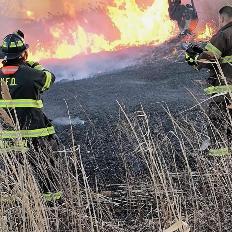 Two firefighters in protective gear work to control a large grass fire, with tall flames and heavy smoke in the background and dry grass in the foreground.