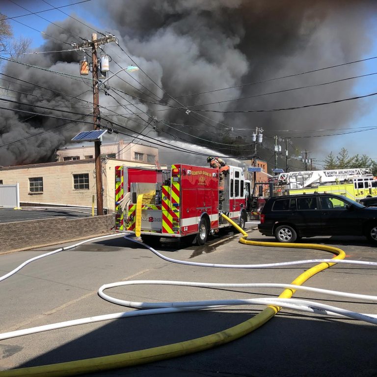A large fire engulfs a building, sending thick black smoke into the sky. Fire trucks and hoses are on the street, and firefighters work to control the blaze. A black SUV is parked nearby.