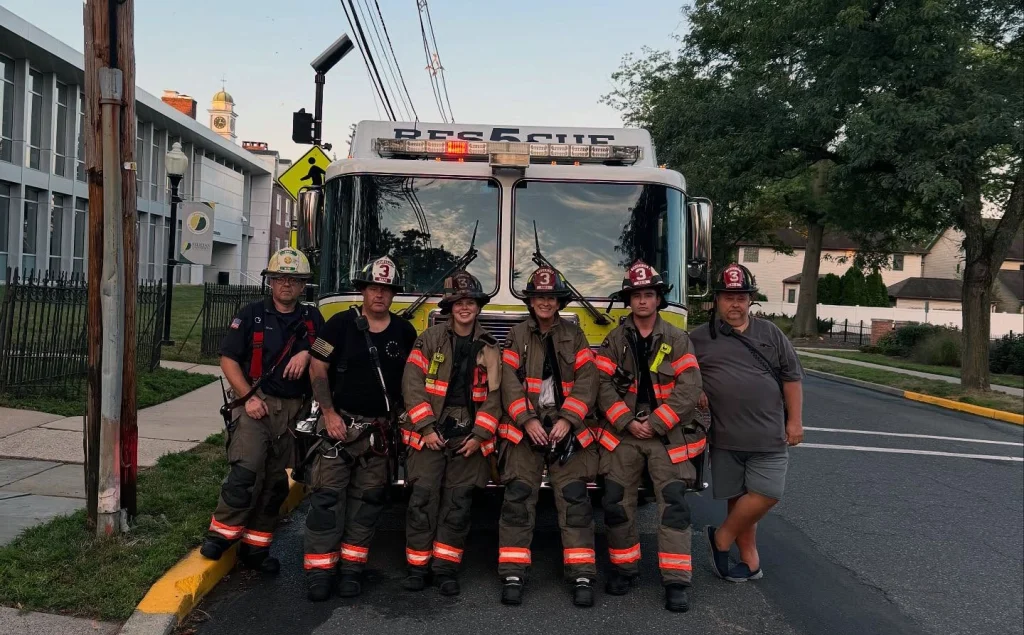 Six people, five in firefighter uniforms and one in casual clothing, stand in front of a rescue fire truck parked on a street. Buildings, trees, and a crosswalk sign are visible in the background.