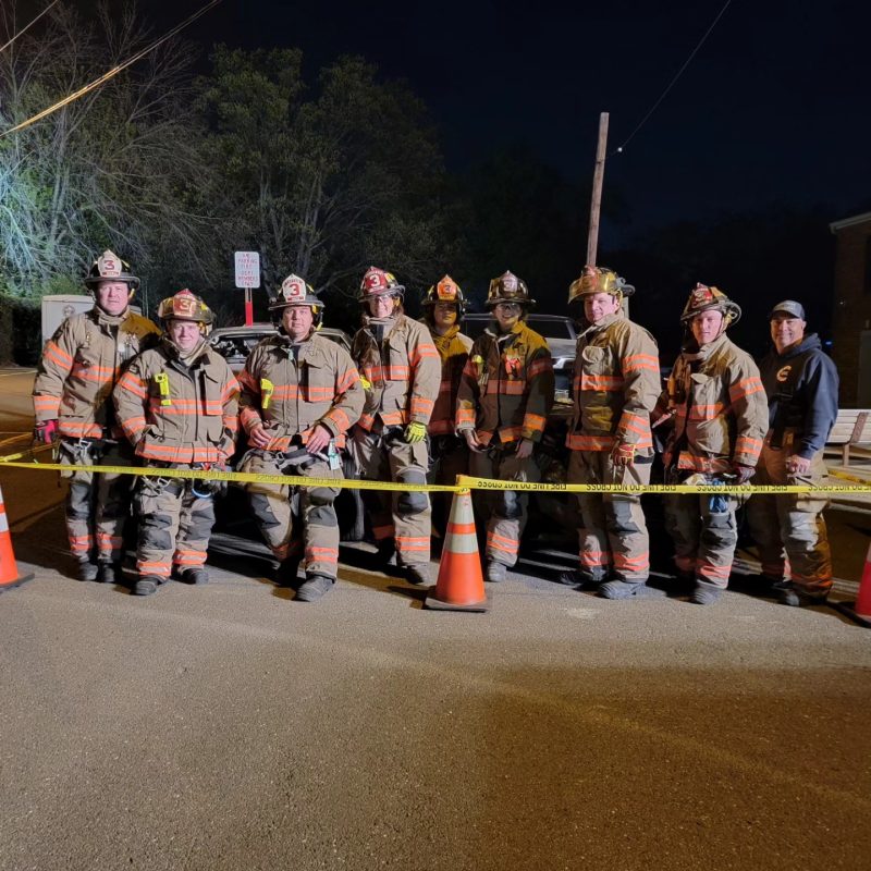 A group of firefighters in full gear stands together at night behind yellow caution tape and orange cones on a street, with trees and a building in the background.