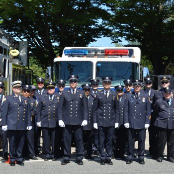 A group of uniformed firefighters stands in front of two fire trucks, one of them holding an American flag. Trees provide shade in the background on a sunny day.