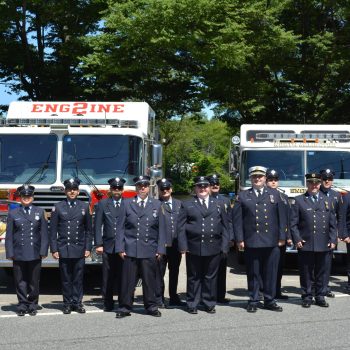 A group of firefighters in uniform stand in front of two fire trucks labeled "Engine 2" during the daytime, with trees and parked vehicles in the background.