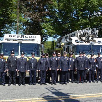 A group of firefighters in dress uniforms stand in a row in front of rescue and fire trucks parked on a sunny street, with trees in the background.