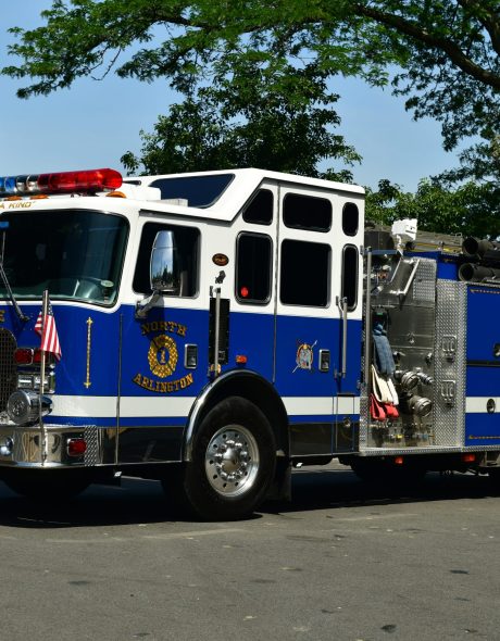 A blue and white fire truck with American flags on the front and sides is parked on a street, surrounded by green trees. The truck is labeled "North Branford" and has "BIG BLUE" on its front license plate.