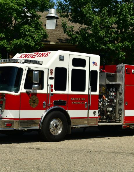 A red and white fire engine labeled "Schuyler Engine Co." is parked in a lot near a building and trees on a sunny day.